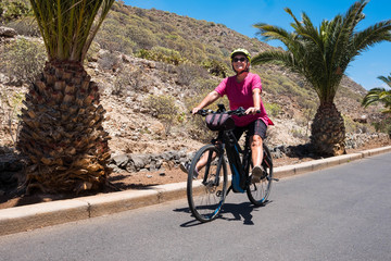 Fototapeta premium Smiles and happiness of a senior lady bicycling with an e bike. Blue sky and palm trees on background. Tropical climate