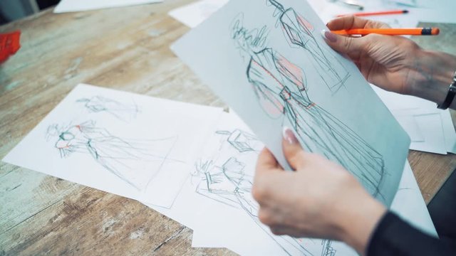 Process of drawing sketches of a model dress on papers by a female tailor. Professional designer draws illustrations for future dresses with a pencil on the table in atelier.