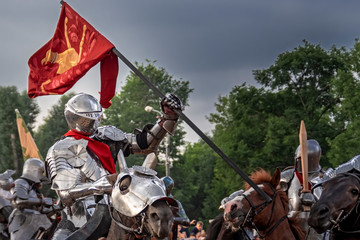Knight on horseback in medieval steel armor with flag in battle.