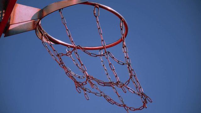 The orange and black basketball ball twice fall through the basket with metal chain net on sunny summer day close up on background of clear blue sky. Concept of modern urban playing field.