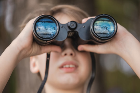 Closeup portrait of cute curious kid exploring nature using old vintage binoculars. Beautiful blue sky, river and nature reflected in glass of lenses. Horizontal color photography.