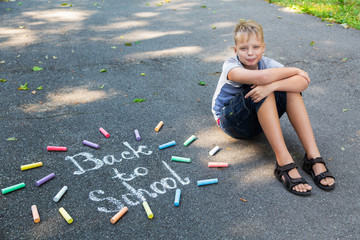 Phrase Back to school drawn with white chalk on grey surface of sidewalk. Portrait of happy smiling white kid sitting on ground. Horizontal color photography.