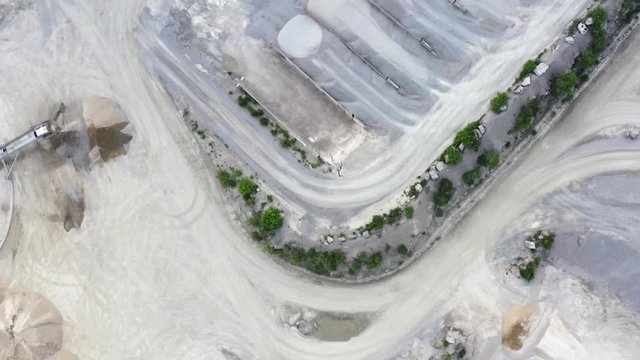 Top down forward aerial of two front loaders working in a stone quarry. Sorted piles of gravel and an industrial outdoor machine pass by on the ground. Location Blekinge in Sweden.