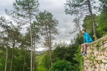 Young man sitting alone on stones wall and looking at green nature view with forest on background. Girona, Catalonia, Spain. Man in nature concept.