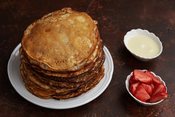 Homemade pancakes or crepes served with condensed milk and strawberries on a brown background. Top view.