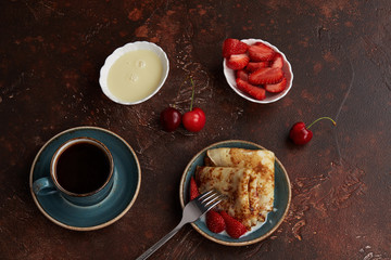 Espresso Cup and homemade pancakes or crepes served with condensed milk, strawberries and cherries on a brown background. Top view.