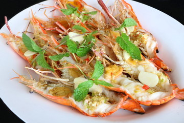 Side view, close up grilled fresh river shrimp with seafood sauce, slice garlic and mint leaves in white plate on black background is famous food in Thailand look delicious.