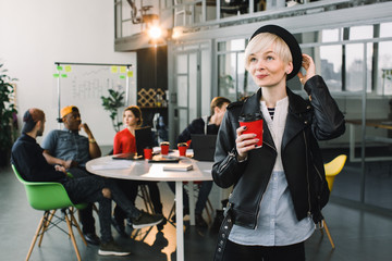 Beautiful Woman in black hat and jacket With red cup of Coffee standing in modern Office . Portrait Of Successful Business Woman Holding Cup Of Hot Drink In Hand
