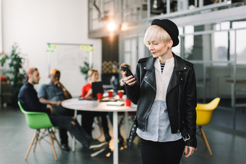 Caucasian pretty business woman in hat and jacket using phone. successful European business woman, talking on the phone, standing on background of colleagues on meeting in modern loft office