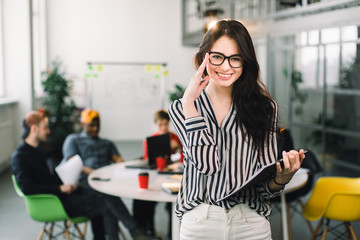 Beautiful trendy brunette business woman in glasses with folder of documents in her hands indoors. Team of young workers discussing on the background