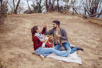 Attractive smiling multicultural couple dressed casual sitting on blanket at picnic, eating sandwiches and giving high five. Autumn season.