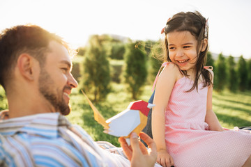 Happy child playing with her father with a bird toy sitting on the green grass outdoors. Young dad smiling and enjoying at day out with his daughter in the park. Daddy and little girl shares love.