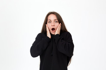Studio shot of a beautiful young woman in a black sweatshirt expressing emotion of surprise while standing over white background and shrugging her shoulders.