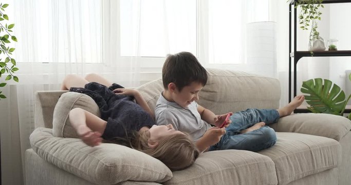Girl lying upside down on sofa while her brother is playing game on mobile phone