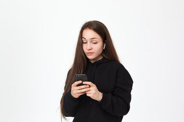 Fototapeta premium Studio shot of a beautiful young brunette woman with wireless headset looking with skeptical emotion at the phone while standing over white background. Girl wears black hoodie.