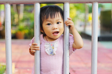 Obraz premium Happy baby child girl standing and holding a steel fence at the playground. She smiling and playing a toy.
