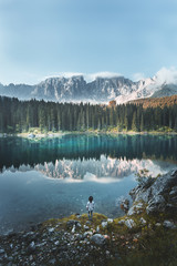 Rear view of a woman standing by the Karersee lake or Lago di Carezza with reflection of mountains in the Dolomites, South Tyrol, Italy.