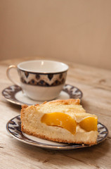 Slice of peach apricot pie or cheese sour cream cake on bright saucer with tea cup on wooden table