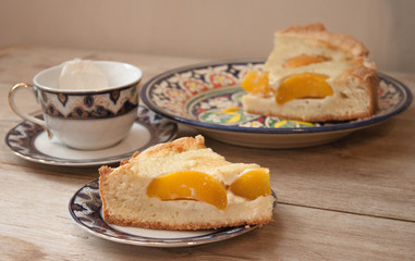 Slice of peach apricot pie or cheese sour cream cake on bright saucer with tea cup on wooden table