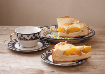 Slice of peach apricot pie or cheese sour cream cake on bright saucer with tea cup on wooden table