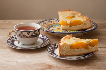 Slice of peach apricot pie or cheese sour cream cake on bright saucer with tea cup on wooden table