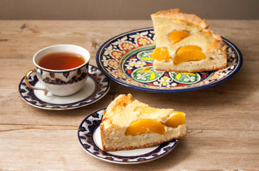 Slice of peach apricot pie or cheese sour cream cake on bright saucer with tea cup on wooden table