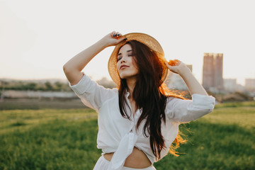 Obraz premium Portrait of beautiful carefree long hair asian girl in white clothes and straw hat enjoys life in field at sunset. Sensitivity to nature concept