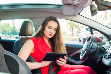 Portrait of serious business lady, caucasian young woman driver in red summer suit setting up the route on a navigator while sitting behind the wheel car. Side view. Selective focus, copy space.