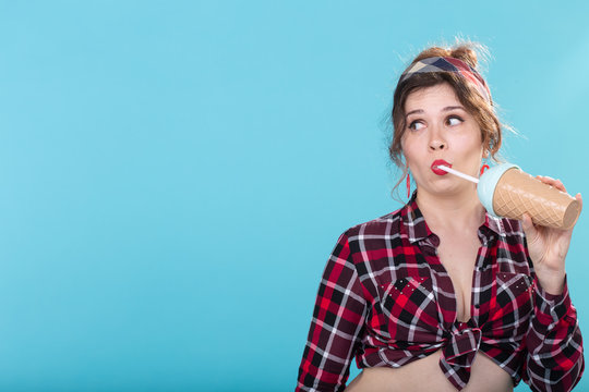 Cute charming young woman in retro clothes is drinking a cocktail from a straw posing against a blue background with copy space and looking to the right. Concept of links and information.