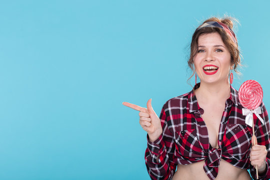 Smiling beautiful young girl in a plaid shirt with a lollipop in her hands posing on a blue background and shows a finger to the right. Concept information and subscription link. Copy space