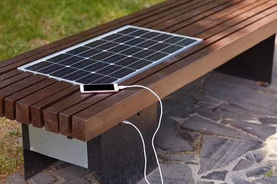Outdoor shot of bench with smart phone being charged via USB in city park. Wooden bench with solar panel for people. Modern technology, altenative electricity, ecology concept.