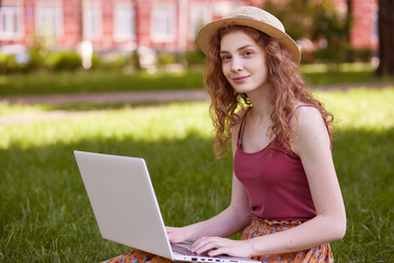 Horizontal shot of pretty young woman sitting on green grass in park with portable computer on legs during summer day, using wireless INternet for online working, female wearing t shirt and straw hat.