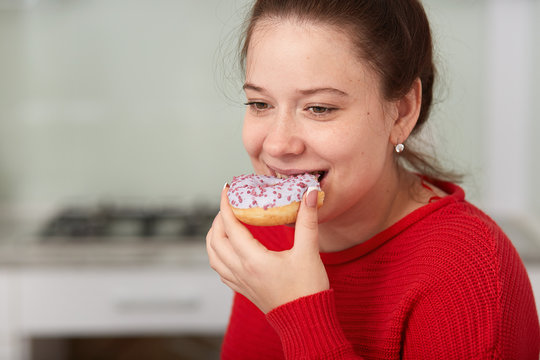 Close Up Portrait Of Young Happy Woman Eating Cake While Sitting At Table In Kitchen, Lady Wearing Red Casual Sweater, Has Happy Facial Expression, Looking Down. Unhealthy Eating And Lifestyle Concept