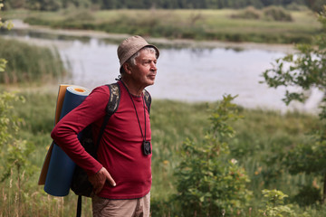 Outdoor shot of eldery man with backpack and rug, senior male poing in open air with hands on hips, decides where to go, wearing red casual sweater and cap. Backpacking and trekking concept.
