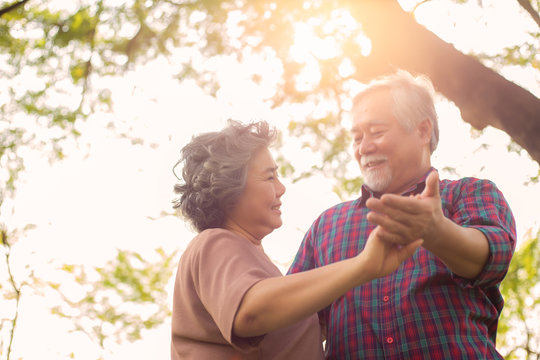 Old woman or wife dancing with old man or husband at park under tree in morning. Grandmother and grandfather feel happiness at the wonderful moment time with smiley faces. Senior couple is romantic