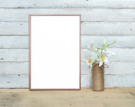 Rose Gold vertical A4 Frame mockup near a bouquet of lilies stands on a wooden table on a painted white wooden background. Rustic style, simple beauty. 3 render.