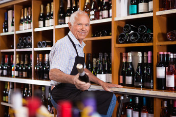 Confident elderly male owner of wine shop taking wine bottle from shelf rack and proffering to buy
