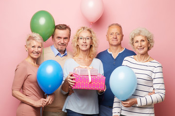 Smiling mature lady and her friends holding colorful balloons standing against pink wall and...