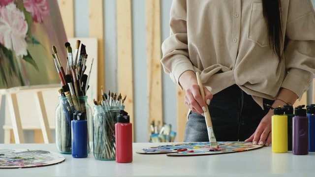 The hands of a young woman professional artist working in her workshop at the table with a set of brushes and a palette of watercolors. Concept of the artist's work