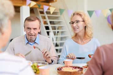 Positive mature friends in casual sweaters sitting at table and eating food while discussing news at dinner party