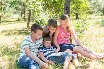 Naklejka na ściany i meble Children learn in nature, four children read a book in the park in the open air. Pupils are preparing for school.