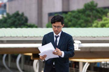 Young Asian business man in suit analyzing charts or paperwork in hands outdoors.