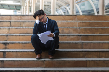 Depressed stressed young Asian business man in suit with hands on head sitting on stairs. Unemployment and layoff concept.