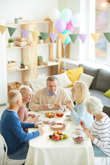 Content senior man with beard sitting at dining table with treats and drinking tea with friends at home