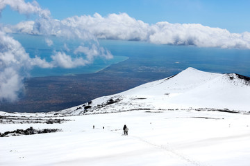 Amazing view from Mount Etna photographed with hikers going down on the snow and sea coast in the background. Magnificent clouds close to the top of the mountain. Etna, Sicily, Italy