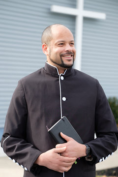 A african-american Male Reverend stands in front of a church with a bible and a black robe