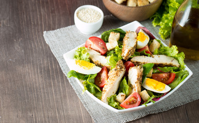 Fresh Caesar salad made of tomato, ruccola, chicken breast, eggs, arugula, crackers and spices. Organic ingredients in a white, transparent bowl on wooden background