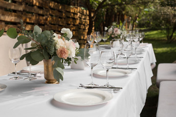 Wedding table setting decorated with fresh flowers in a brass vase. Wedding floristry. Banquet table for guests outdoors with a view of green nature. Bouquet with roses, eustoma and eucalyptus leaves