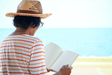 An elderly Asian woman reading a book at the beach by the sea