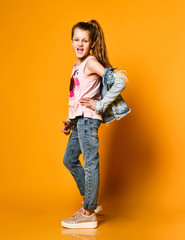 young teen girl model posing on a yellow background in jeans and a jacket, with long hair gathered in a high tail, stands half a turn to the camera.